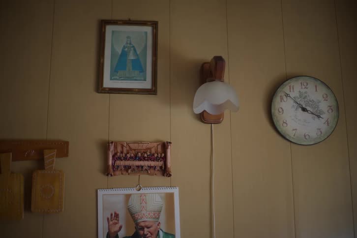 A wall in a home is decorated with Catholic religious icons, including images of Pope John Paul II, the Last Supper, and a vintage clock