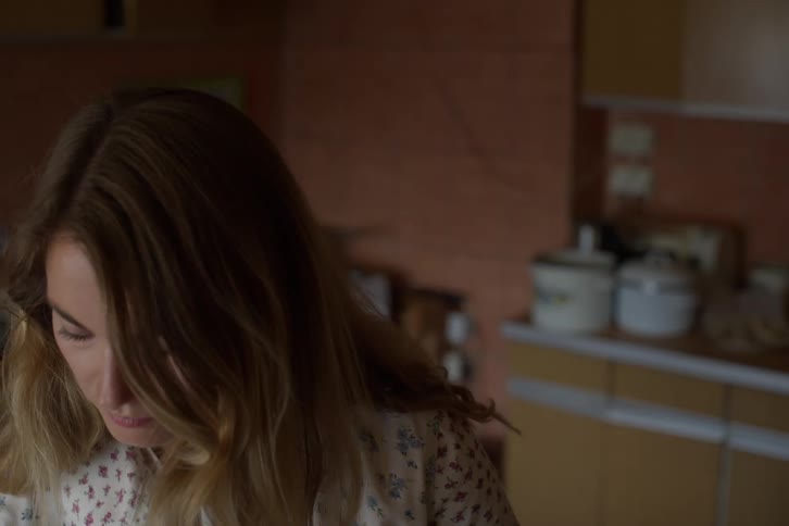 A thoughtful woman with long hair looks down while standing in her dimly lit, vintage kitchen