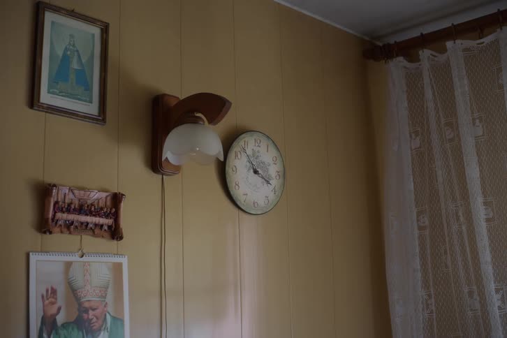 Time passes on a vintage wall clock next to religious pictures and a lamp in an old-fashioned home