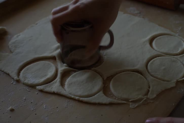A baker's hands use a glass cup to cut out circular shapes from rolled-out dough on a wooden board