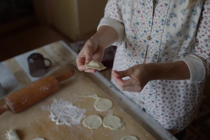 A person's hands meticulously shape a traditional homemade dumpling over a floured wooden board