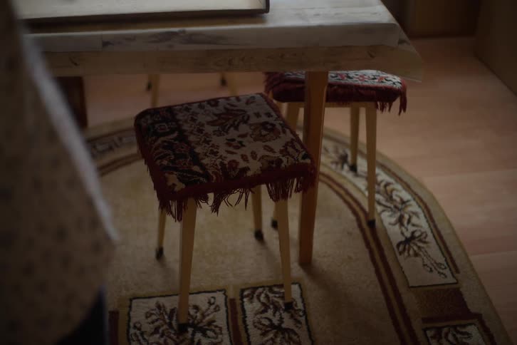 A person stands in front of a simple wooden table with old-fashioned fringed stools in a dimly lit, modest room