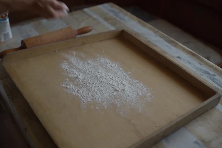 A person's hand dusts a rustic wooden board with white flour in preparation for baking