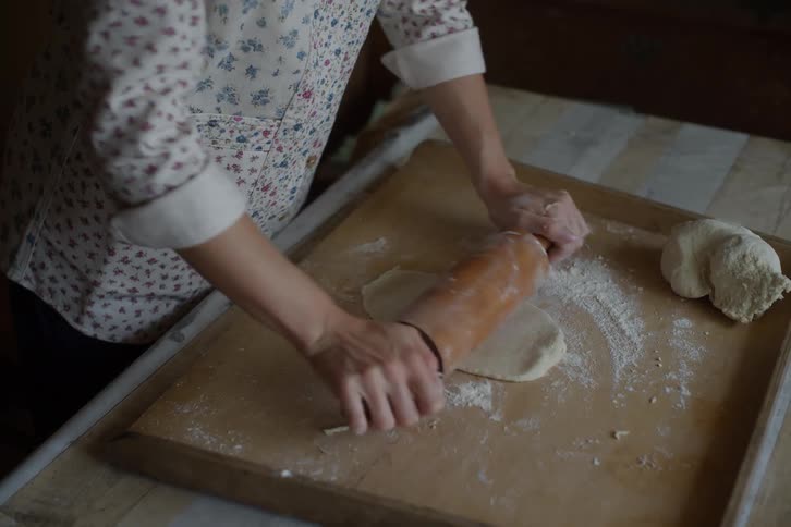 A woman wearing a floral shirt kneads and rolls out fresh dough on a floured wooden board with a rolling pin