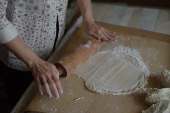 A woman's hands roll out fresh dough with a wooden rolling pin on a floured kitchen board, preparing for baking