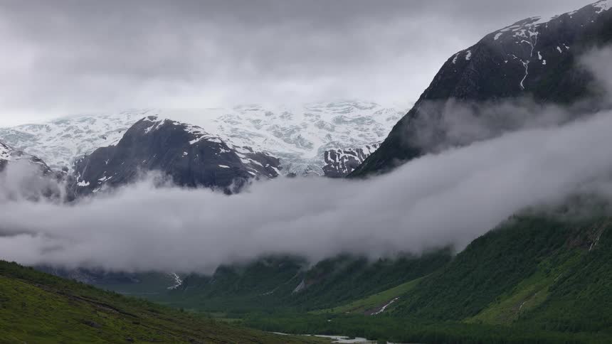A stunning view of a snow-covered mountain glacier with thick fog rolling through a lush green valley on an overcast day