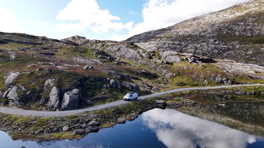 Aerial view of a white camper van parked on a remote mountain road next to a calm, reflective lake in Norway's wilderness