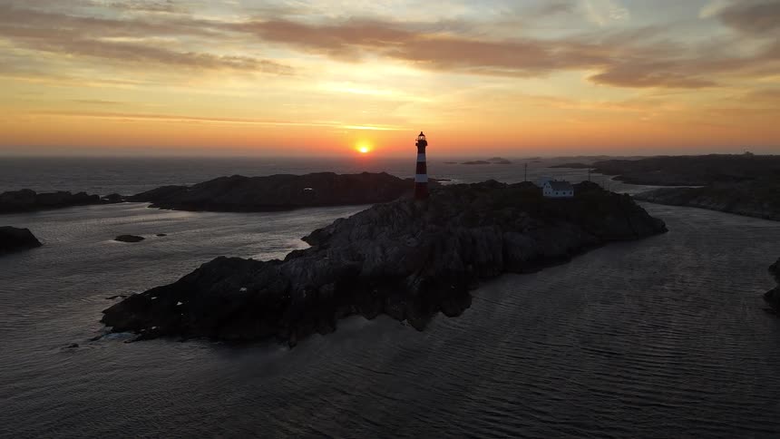 A serene aerial view captures a red and white striped lighthouse standing on a rocky island as the sun sets beautifully over the ocean horizon