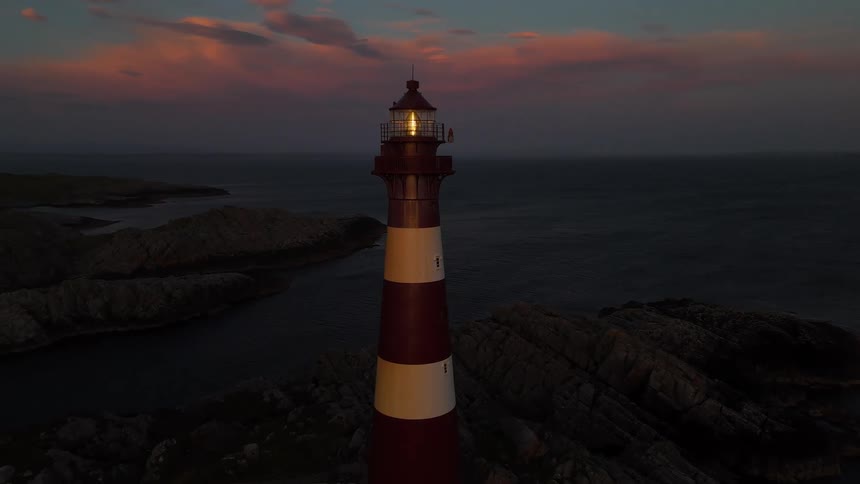 An aerial view pulls back from a classic red and white striped lighthouse as its light shines over the rocky coast at sunset