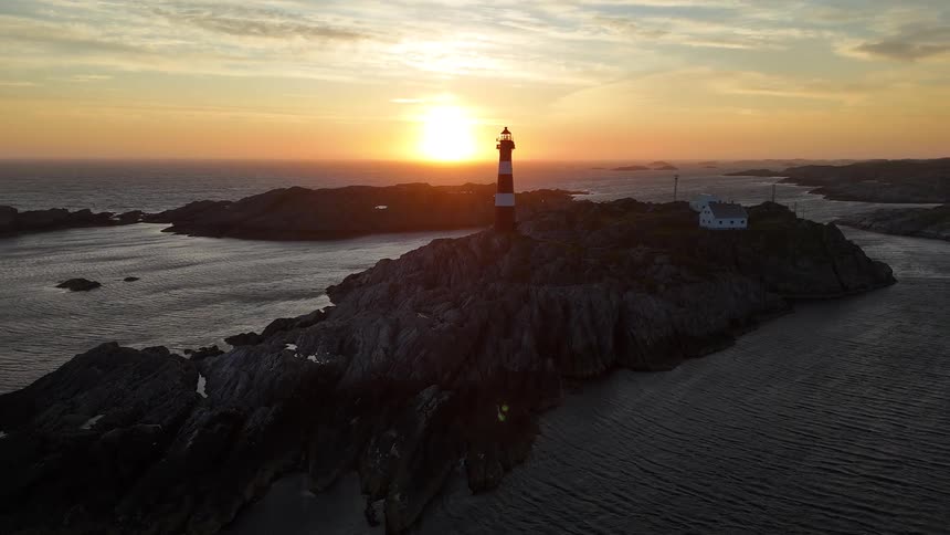 An aerial pullback shot reveals a picturesque red and white lighthouse silhouetted against a brilliant sunset on a remote rocky island