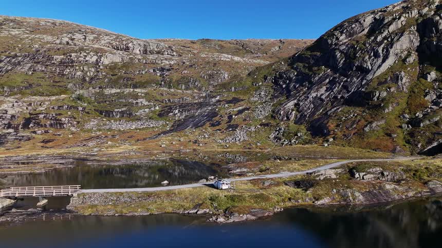 Aerial view pulls back from a white camper van parked on a scenic road next to a tranquil mountain lake in a vast, rocky landscape