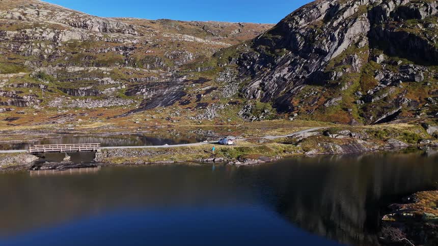 A serene, static shot captures a picturesque mountain landscape with a still lake, a roadside bridge, and people enjoying a peaceful day