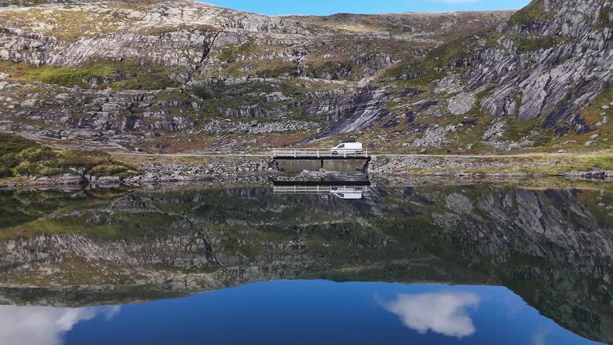 A white camper van drives over a small bridge on a mountain road next to a crystal-clear, reflective lake