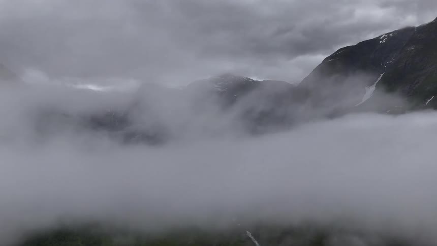 An aerial view slowly rises above thick fog to reveal a stunning, snow-capped mountain range under a moody, grey sky