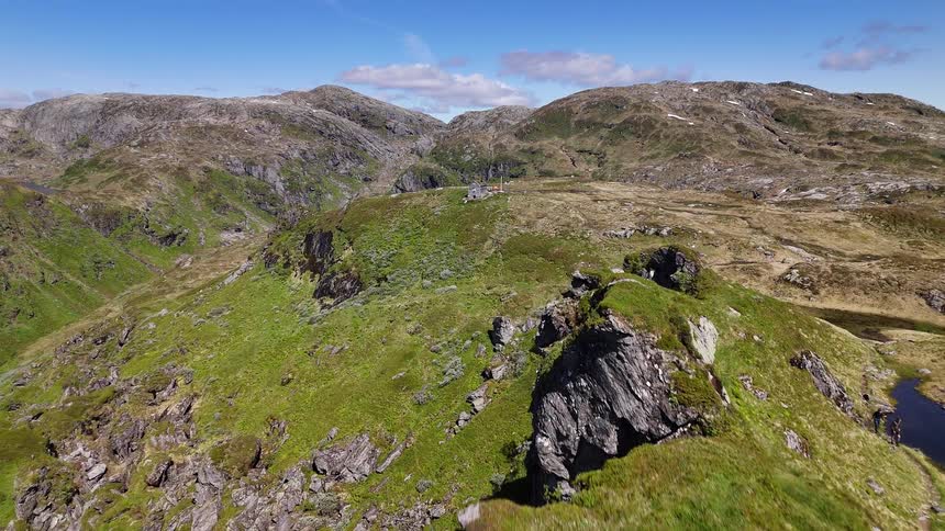 An aerial drone flies over a rugged green mountain ridge, revealing a vast, rocky landscape and a distant cabin