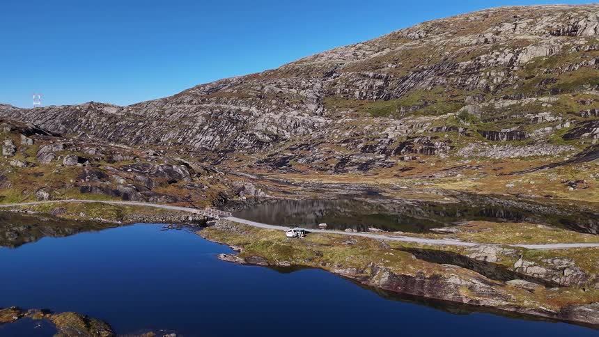 An aerial shot pulls back to reveal a camper van parked on a remote road beside a deep blue lake in a vast and rocky mountain landscape