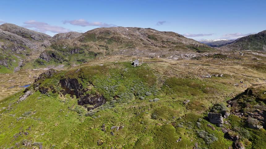 Aerial view slowly pulls back from a remote stone hut on a rugged, green mountainside on a sunny day