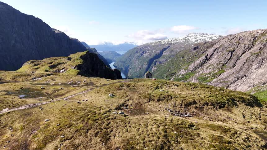 A scenic aerial shot flies over a high mountain plateau with a remote cabin overlooking a beautiful Norwegian fjord