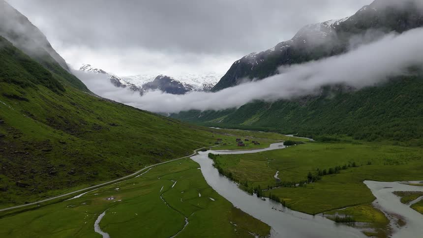 A stunning aerial view of a lush green mountain valley with a winding river, shrouded in dramatic clouds and fog with snow-capped peaks in the distance