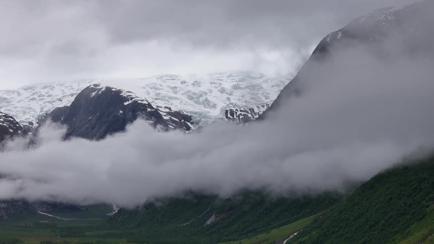 A serene and moody view of a massive glacier nestled in the mountains, with thick fog slowly drifting through the green valley below