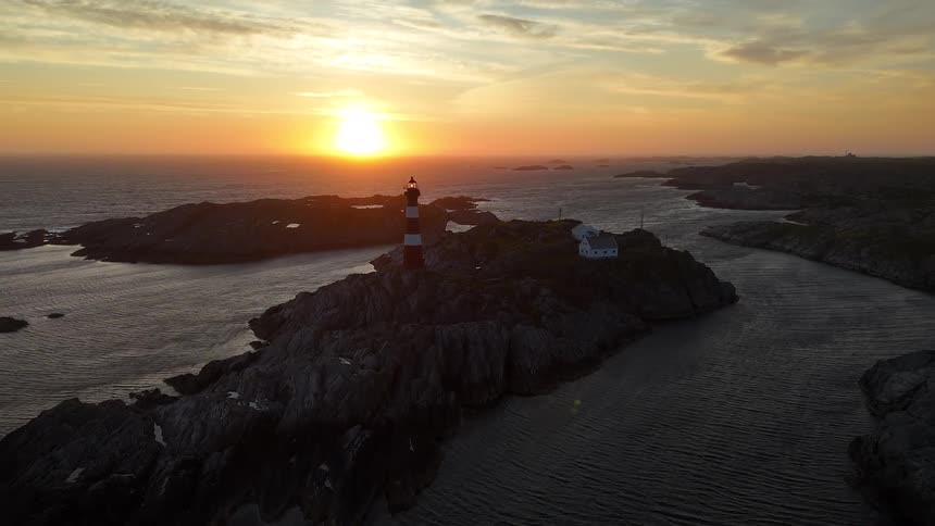 A stunning aerial view pulls back from a classic red and white lighthouse on a rugged, rocky island during a magnificent golden sunset over the sea