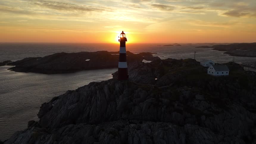 A majestic aerial view reveals a red and white striped lighthouse on a rocky island as the sun sets over the ocean horizon