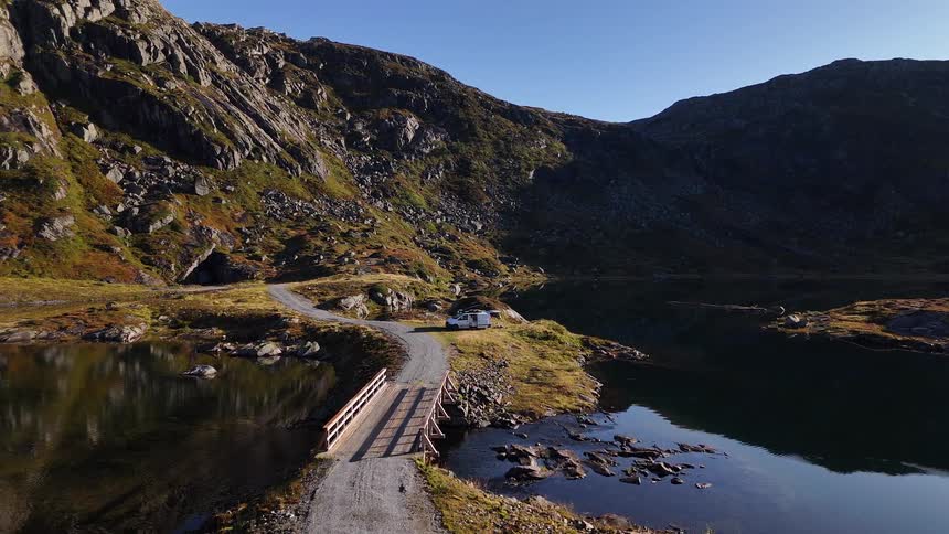 An aerial view shows a camper van parked on a scenic mountain road next to a tranquil lake and small wooden bridge