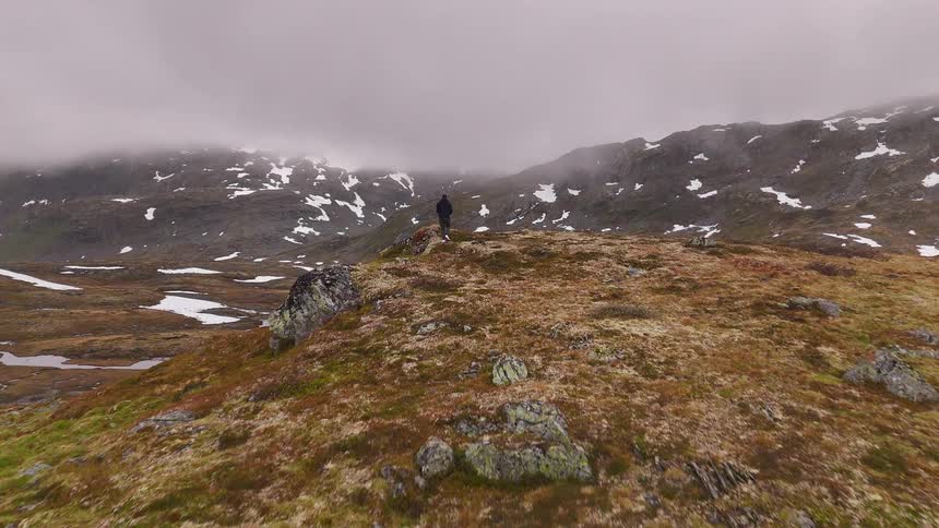 A lone man walks to the edge of a rocky hill to survey the vast, snow-patched, and foggy mountain landscape below