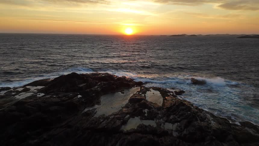 Aerial view flying over a rugged coastline as waves crash and the golden sun sets on the horizon