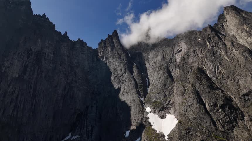 A majestic, jagged mountain range towers towards a blue sky as clouds drift over its snow-dusted peaks