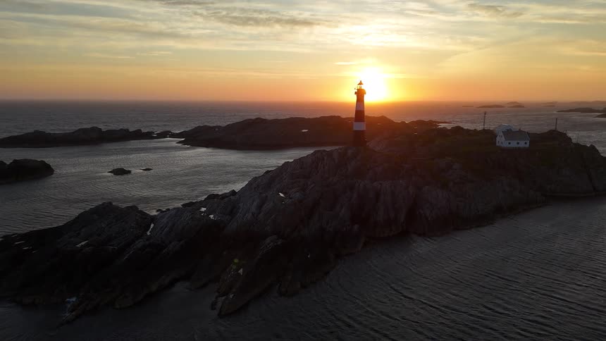 A serene aerial view of a classic red and white striped lighthouse on a rocky island as the sun sets over the calm ocean