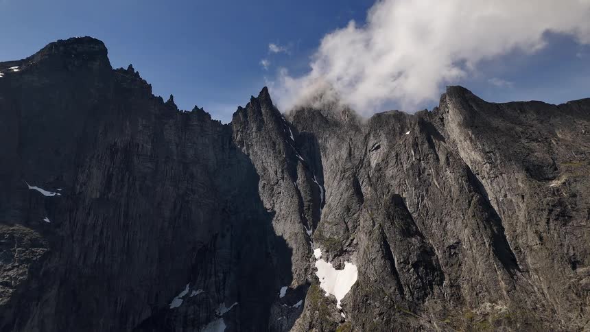 Clouds drift over the rugged and imposing peaks of a mountain range with patches of snow under a clear blue sky