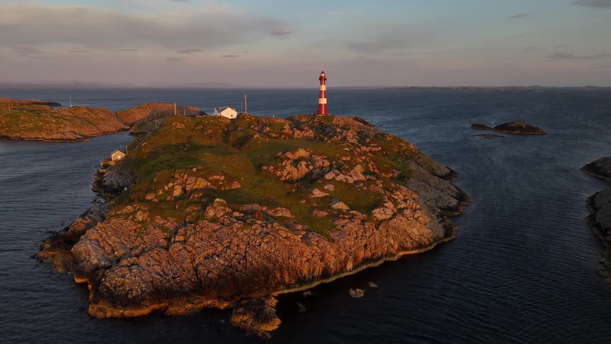A majestic aerial shot slowly approaches a classic red and white lighthouse perched on a rocky island amidst the ocean at sunset