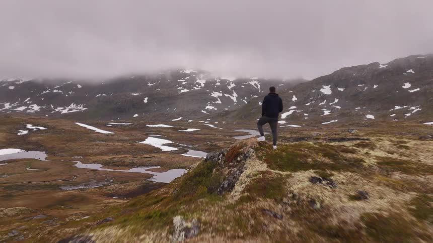 An aerial shot flies towards a lone hiker standing on a hill, admiring the vast and foggy snow-patched mountain landscape