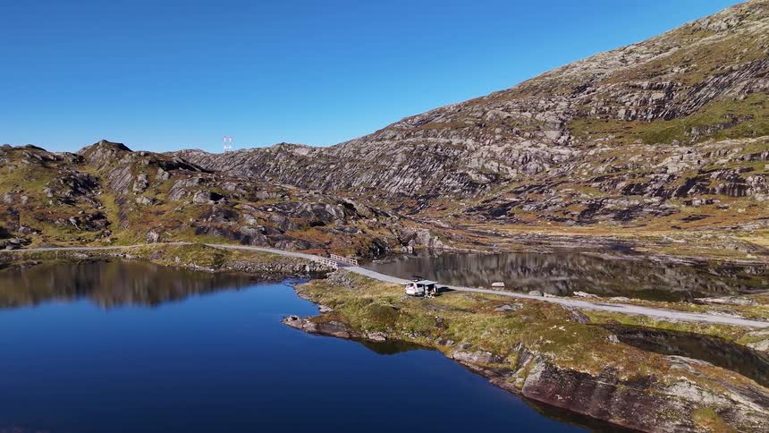 An aerial view shows a camper van parking beside a pristine blue lake in a rugged, mountainous landscape