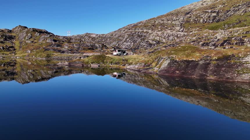 A stunning aerial shot glides over a perfectly calm mountain lake, reflecting the sky and mountains, towards a parked camper van