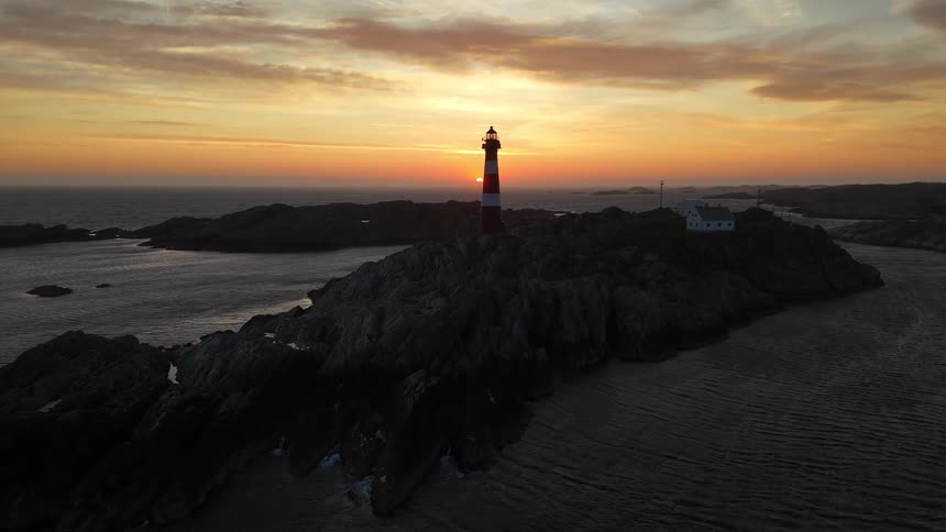 A stunning aerial view of a red and white striped lighthouse silhouetted against a golden sunset on a rocky island coast