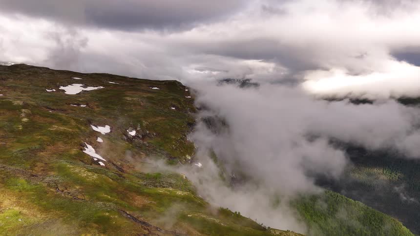 Slow aerial view over a vast, green mountain landscape with snow patches as thick fog drifts up from the valley below under a dramatic, cloudy sky