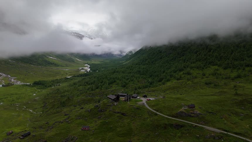 An expansive aerial view reveals a remote mountain lodge nestled in a lush green valley, with snow-capped peaks emerging from the thick, low-hanging clouds