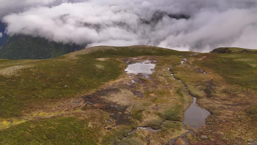 An aerial drone flies over a vast, rugged mountain landscape with patches of snow, revealing a thick blanket of clouds in the valley below