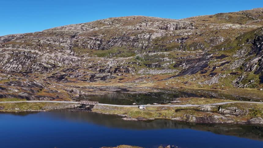 An aerial view shows a camper van parked next to a tranquil blue lake in the rugged, rocky highlands of Norway on a clear day