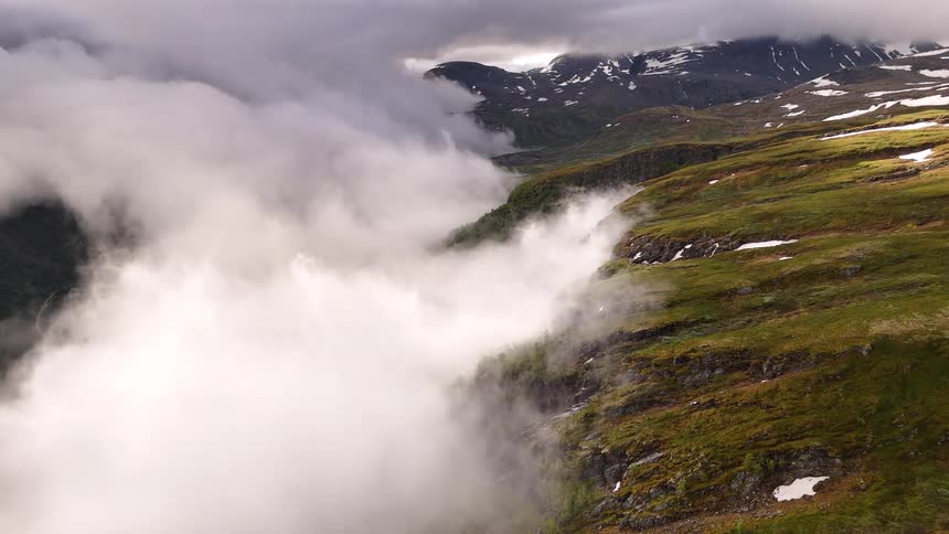 A slow-moving aerial shot captures mist and clouds rolling over a majestic, lush green mountain valley with snow-capped peaks in the distance