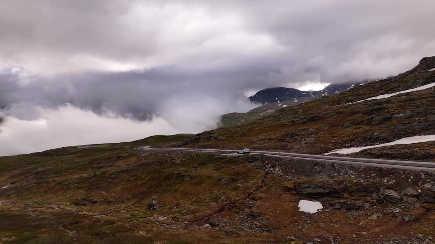 Slowly pulling back aerial shot reveals a solitary car parked on a high mountain road, dwarfed by a vast, snow-patched landscape under a sea of clouds
