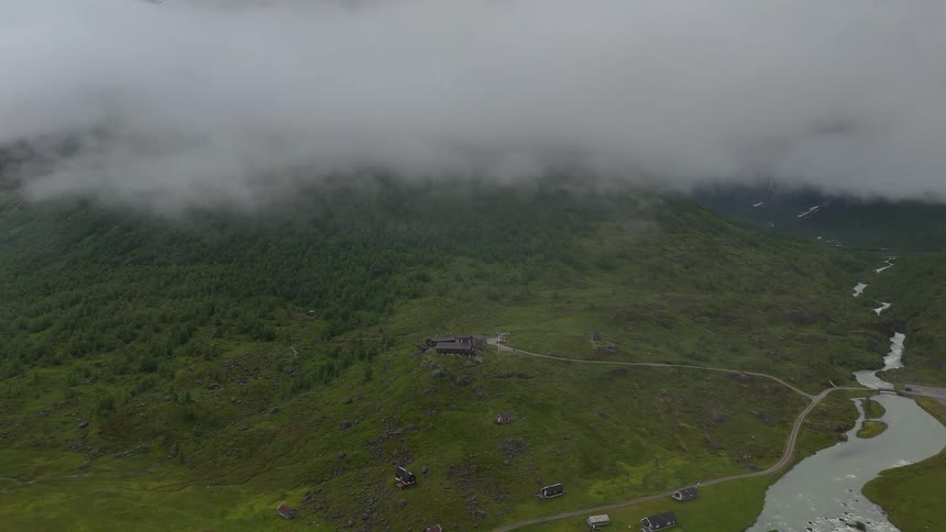 A slow aerial push towards a remote mountain lodge in a lush, green valley shrouded in low-hanging clouds