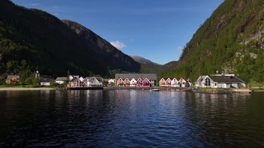 A stunning aerial view pulling back from a colorful village nestled on the tranquil waters of a Norwegian fjord between steep green mountains