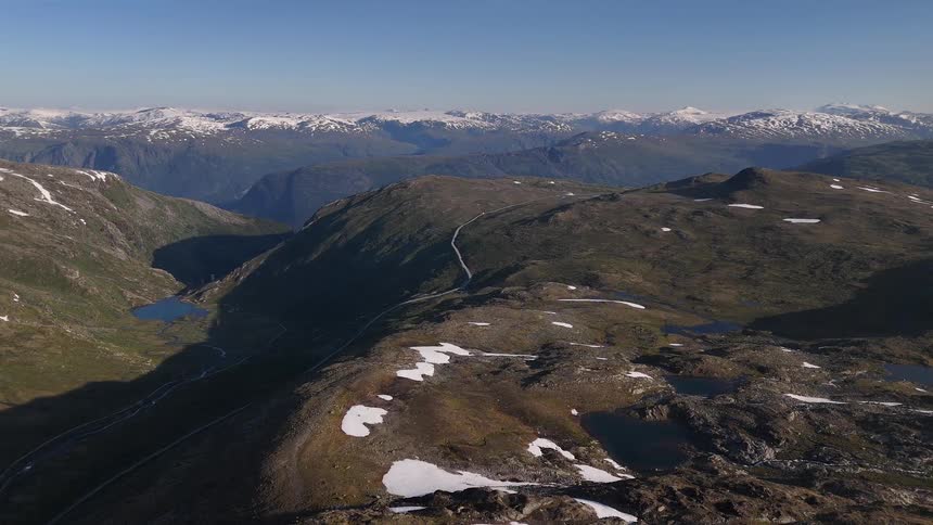 A static aerial view shows a vast, rugged mountain landscape with a winding road, alpine lakes, and distant snow-capped peaks under a clear blue sky