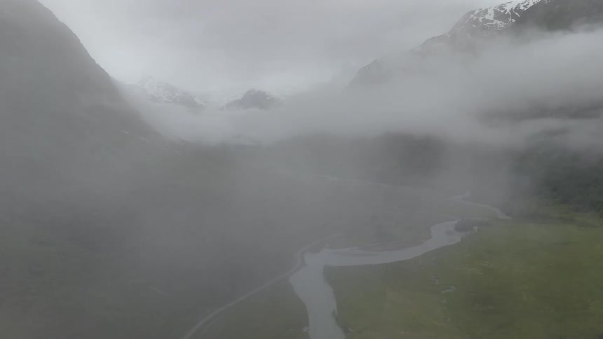 A drone flies backward through thick clouds, revealing a picturesque green valley with a winding river, a remote village, and a distant glacier