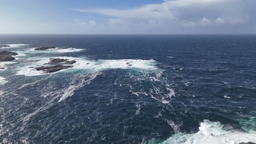 A dramatic, expansive view of the deep blue ocean as powerful waves crash against rocky outcrops on a windy day
