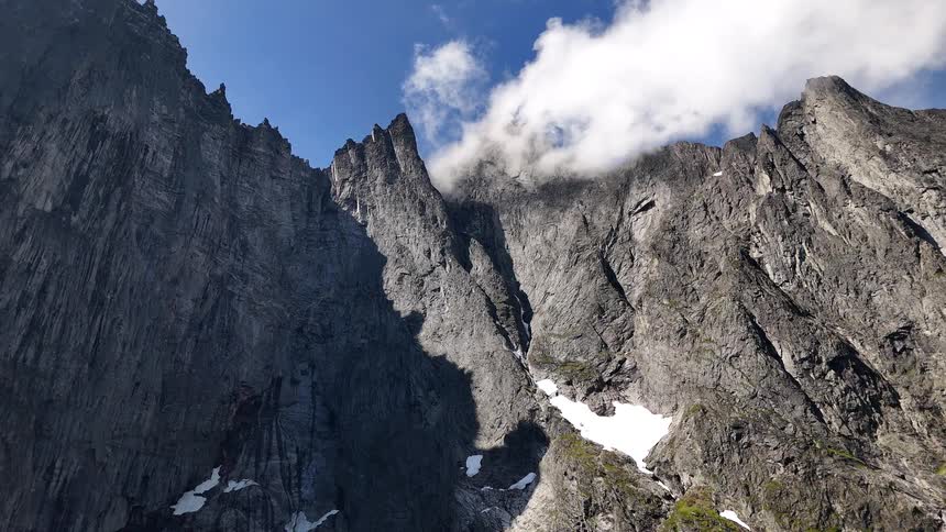 A low-angle view of the towering, craggy Troll Wall mountain range in Norway, with clouds clinging to its peaks against a blue sky