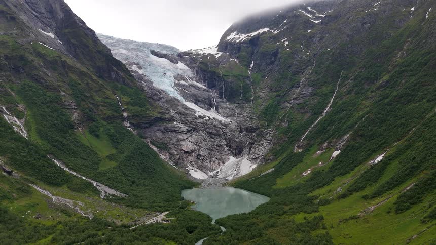 A majestic wide shot of a massive glacier in Norway, with waterfalls from melting ice feeding a turquoise glacial lake in the lush valley below